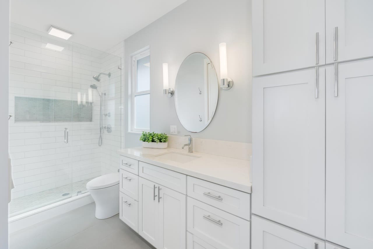 A white bathroom with a round mirror, wall sconces and tall storage